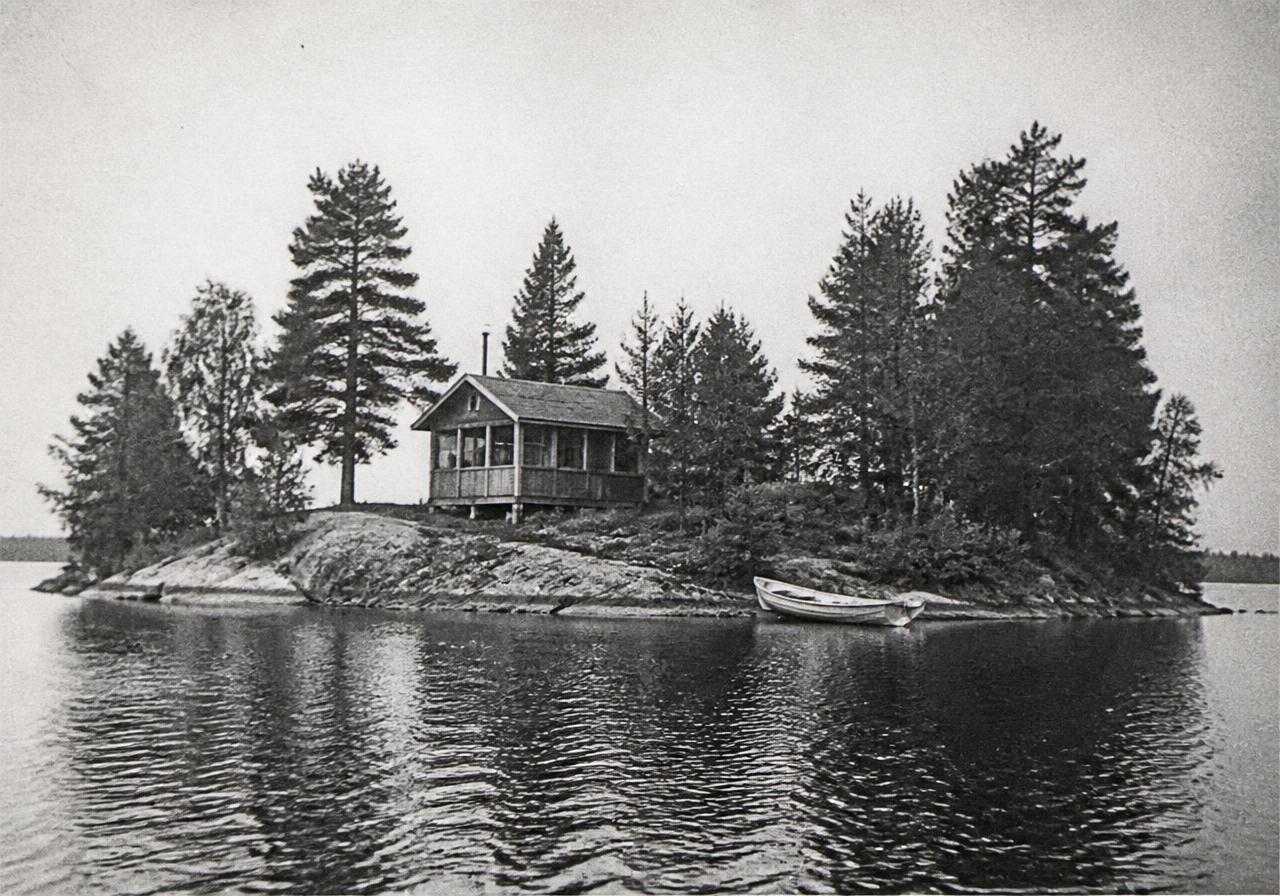 Archival photograph of a small wooded lake island with a modest cottage and rowboat